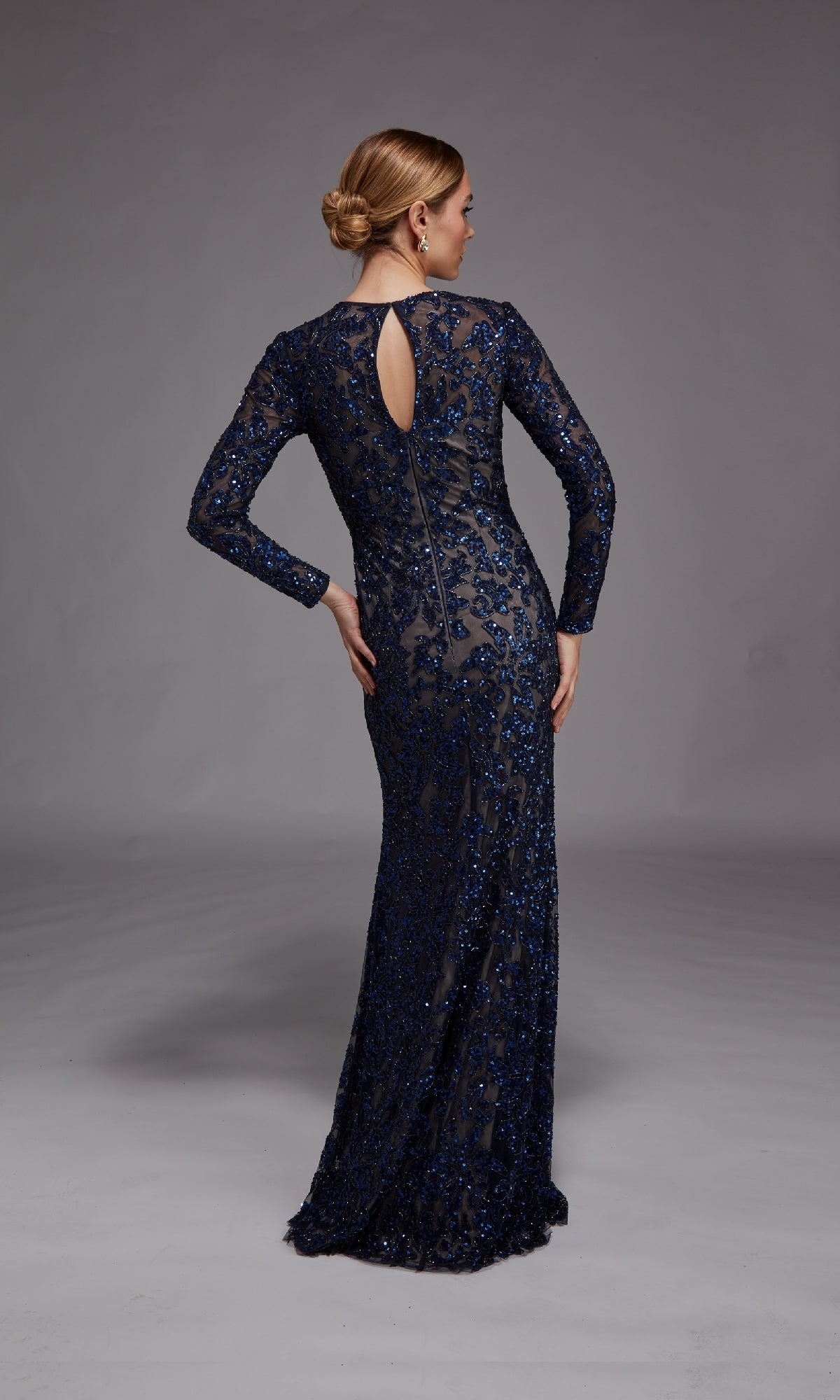 A woman stands with her back to the camera, one hand on her hip, in a floor-length navy blue long-sleeve sequined gown (Long Formal Dress: Alyce 27788) against a plain gray background.